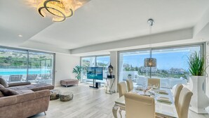 A spacious living area with a large window overlooking a scenic view, a dining area with a table set for four, and a modern chandelier. - 2288 Luxury Penthouse with Private Rooftop Pool (Estepona)