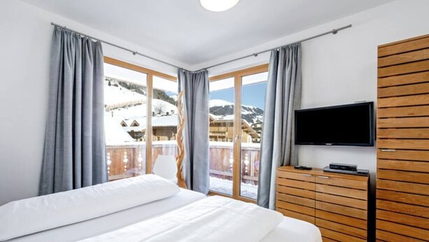 Hotellihuone, jossa on suuri sänky, puinen lipasto, taulutelevisio ja parveke, jolta avautuu näkymä lumihuippuisiin vuoriin.