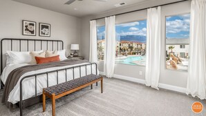 Une chambre avec un lit, un banc, une table de chevet, un ventilateur de plafond et une fenêtre donnant sur une piscine et des bâtiments.