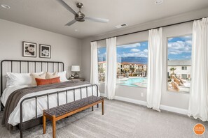 A bedroom with a bed, a bench, a nightstand, a ceiling fan, and a window with a view of a pool and buildings.