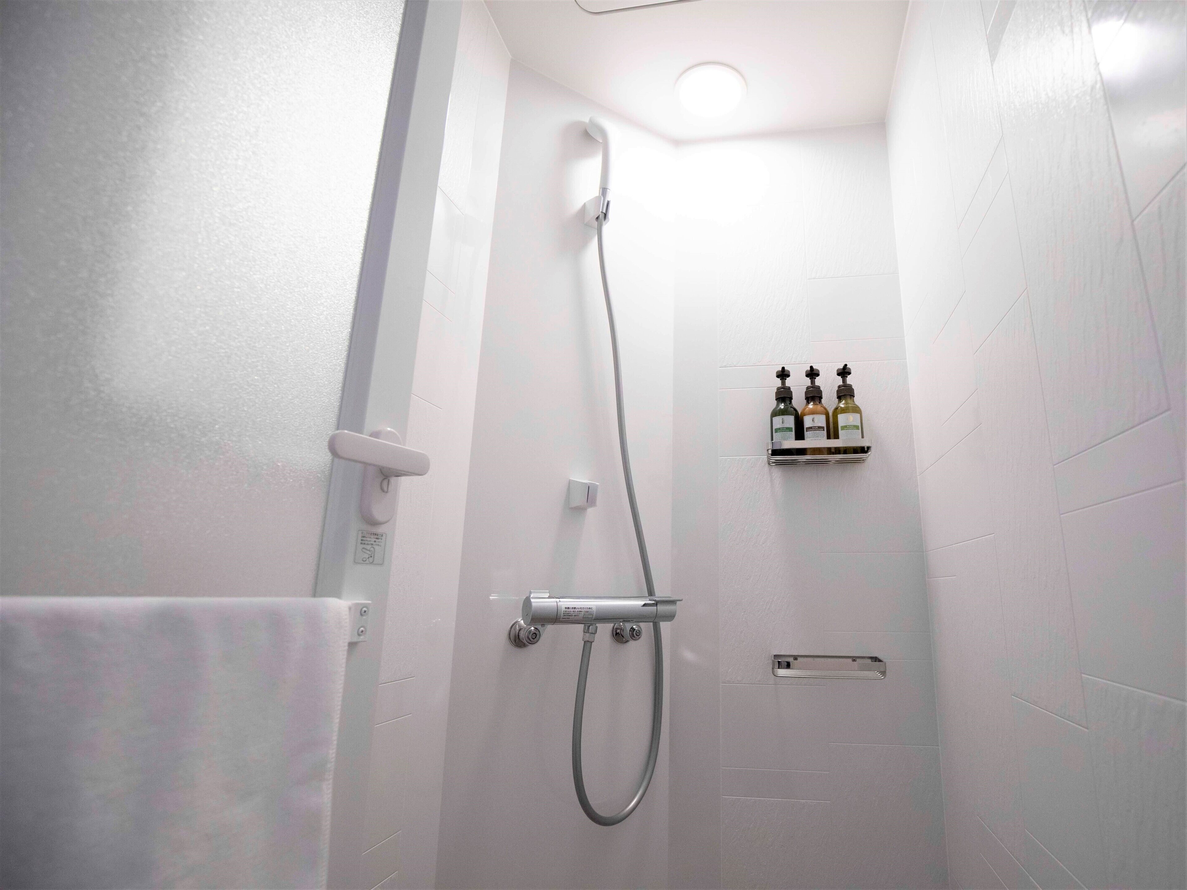 A small, white-walled bathroom with a shower, a towel rack, and a soap dispenser shelf.