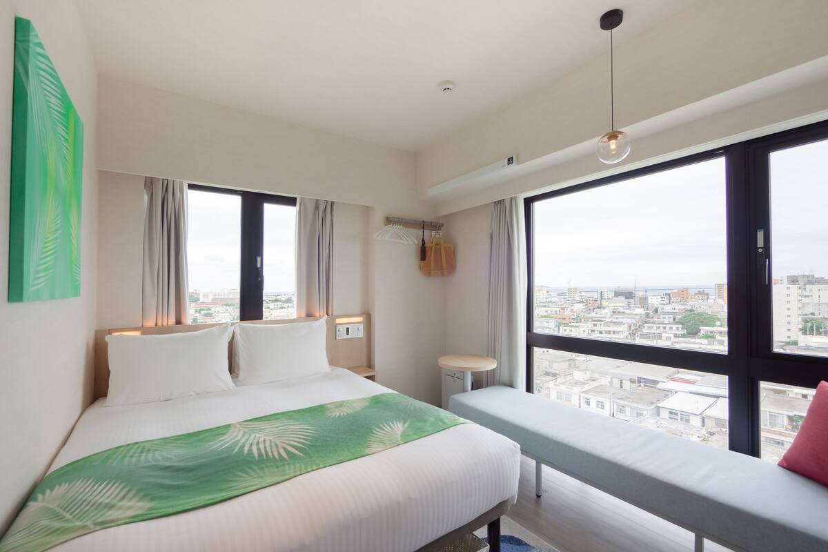 A hotel room with a bed, a bench, a view of the city, and a large window.