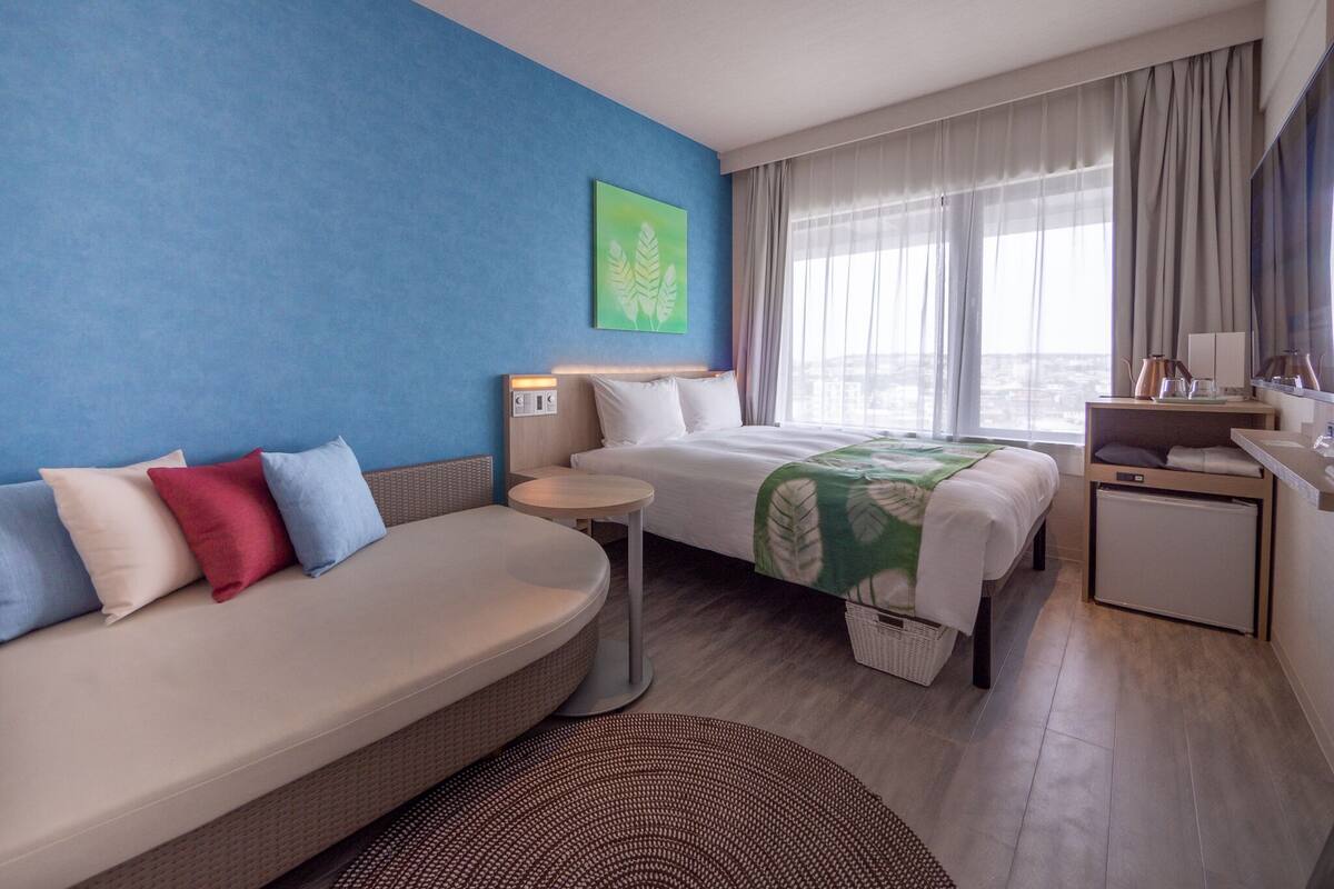 A modern hotel room with a blue accent wall, a bed with a green leaf-patterned bedspread, a sofa with multiple cushions, a small round table, and a view of the city through large windows.