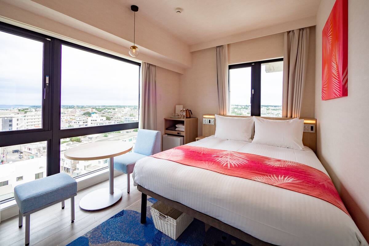 A hotel room with a large bed, a small round table, a bench, and a view of the city through large windows.