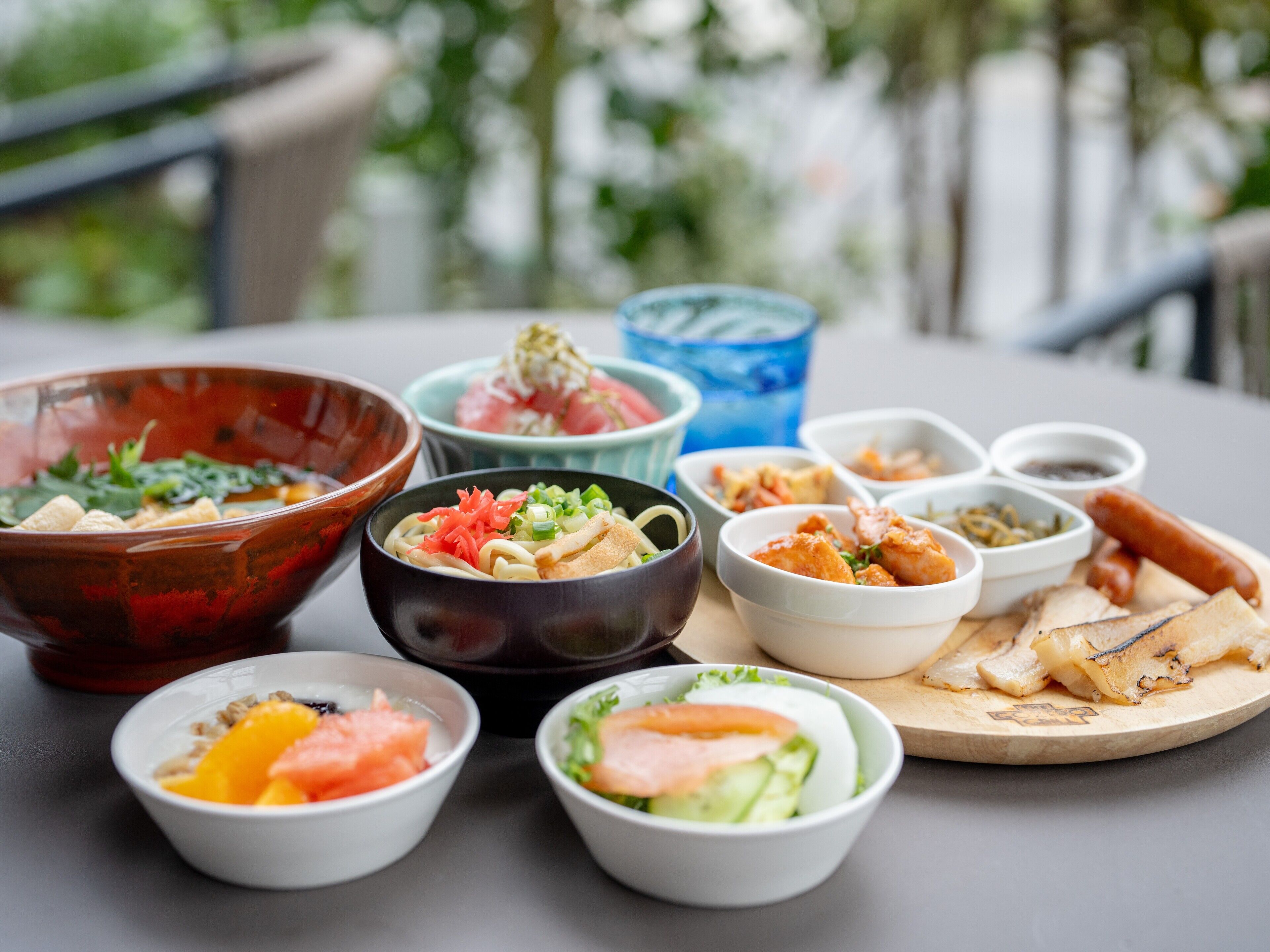 Variedad de platos sobre una mesa, incluyendo tazones de sopa, ensalada y otros alimentos, con una taza azul y una tabla de madera con preparaciones a la parrilla.