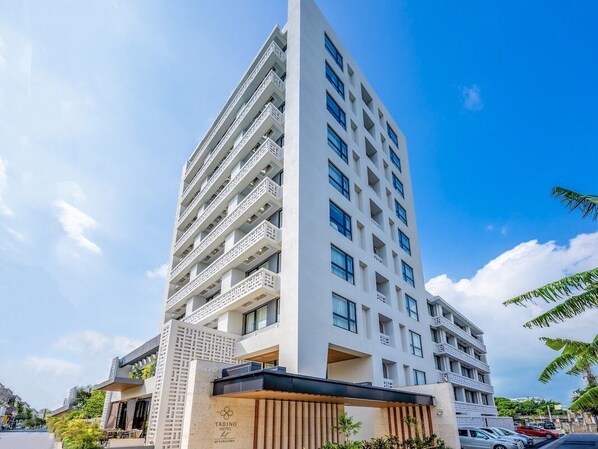 Un hotel moderno de varios pisos con balcones en cada planta, una entrada destacada con fachada de madera y un cielo azul despejado al fondo.