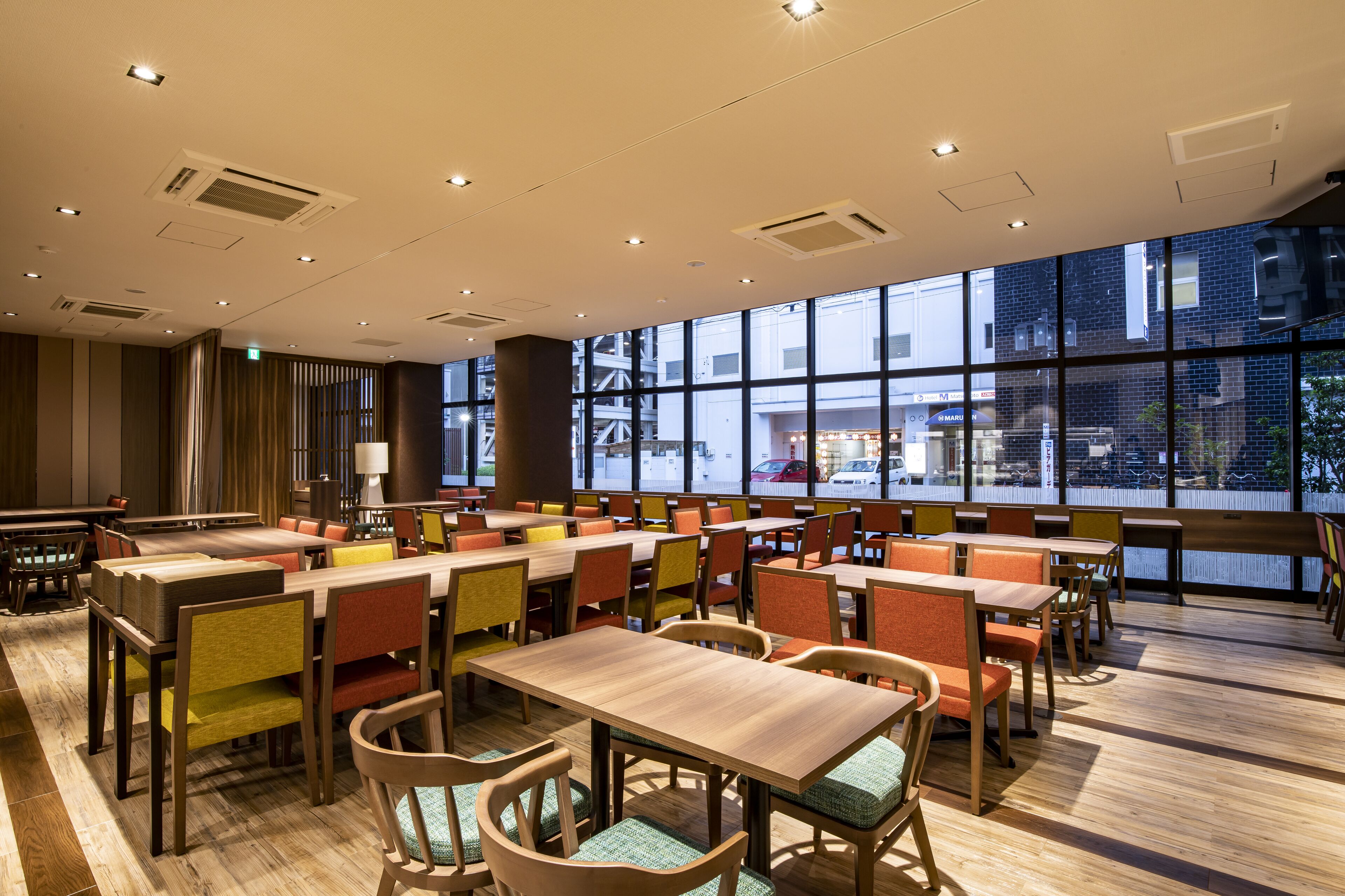 A spacious dining area with multiple tables and chairs, large windows offering a view of the street, and a modern ceiling design with recessed lighting.