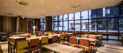 A spacious dining area with multiple tables and chairs, large windows offering a view of the street, and a modern ceiling design with recessed lighting.