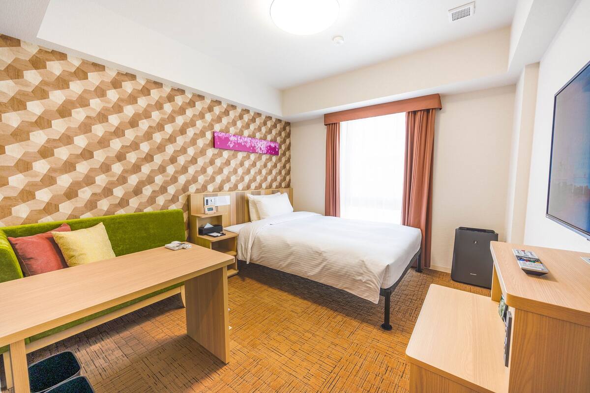 A hotel room with a bed, a green sofa, a wooden desk, a television, and a patterned wall.