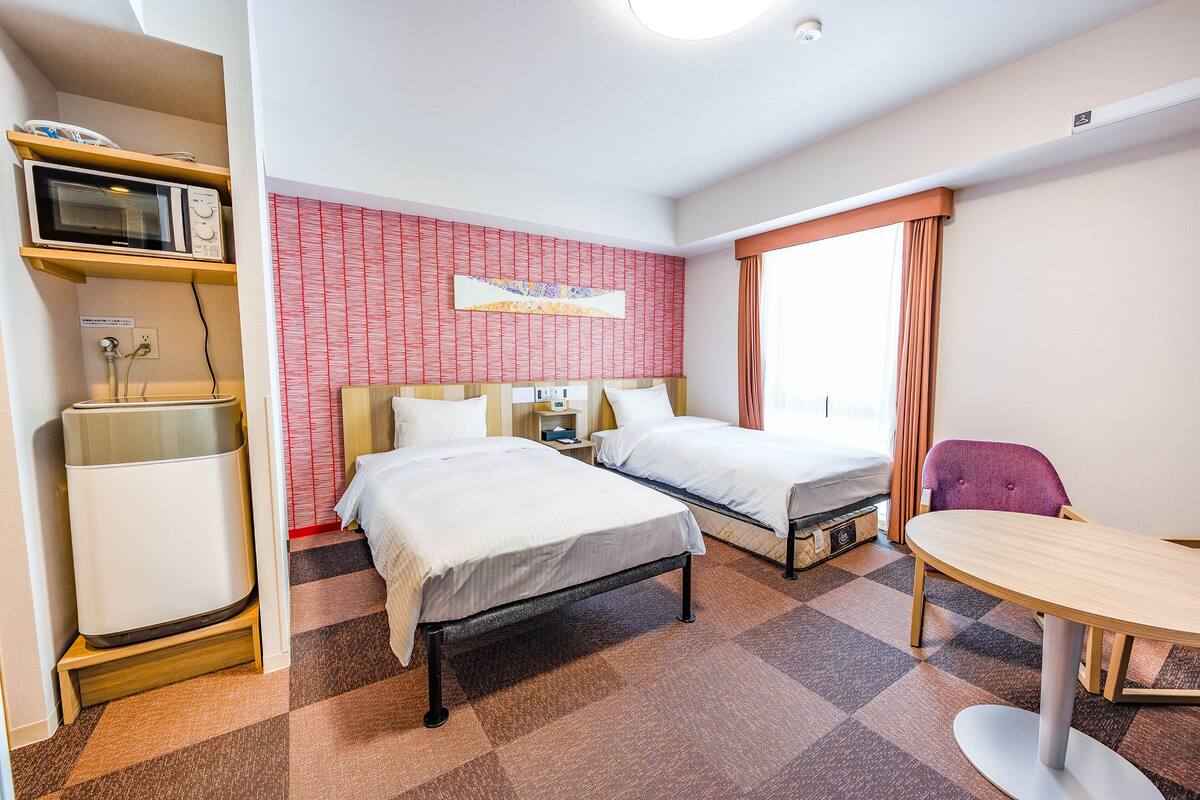 A hotel room with two beds, a microwave, a small table, and a chair.