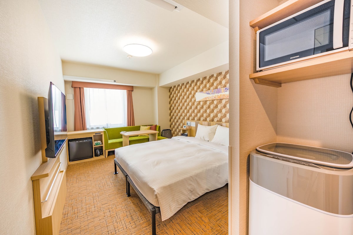 A hotel room with a bed, a TV, a microwave, a small kitchenette, and a window with curtains.