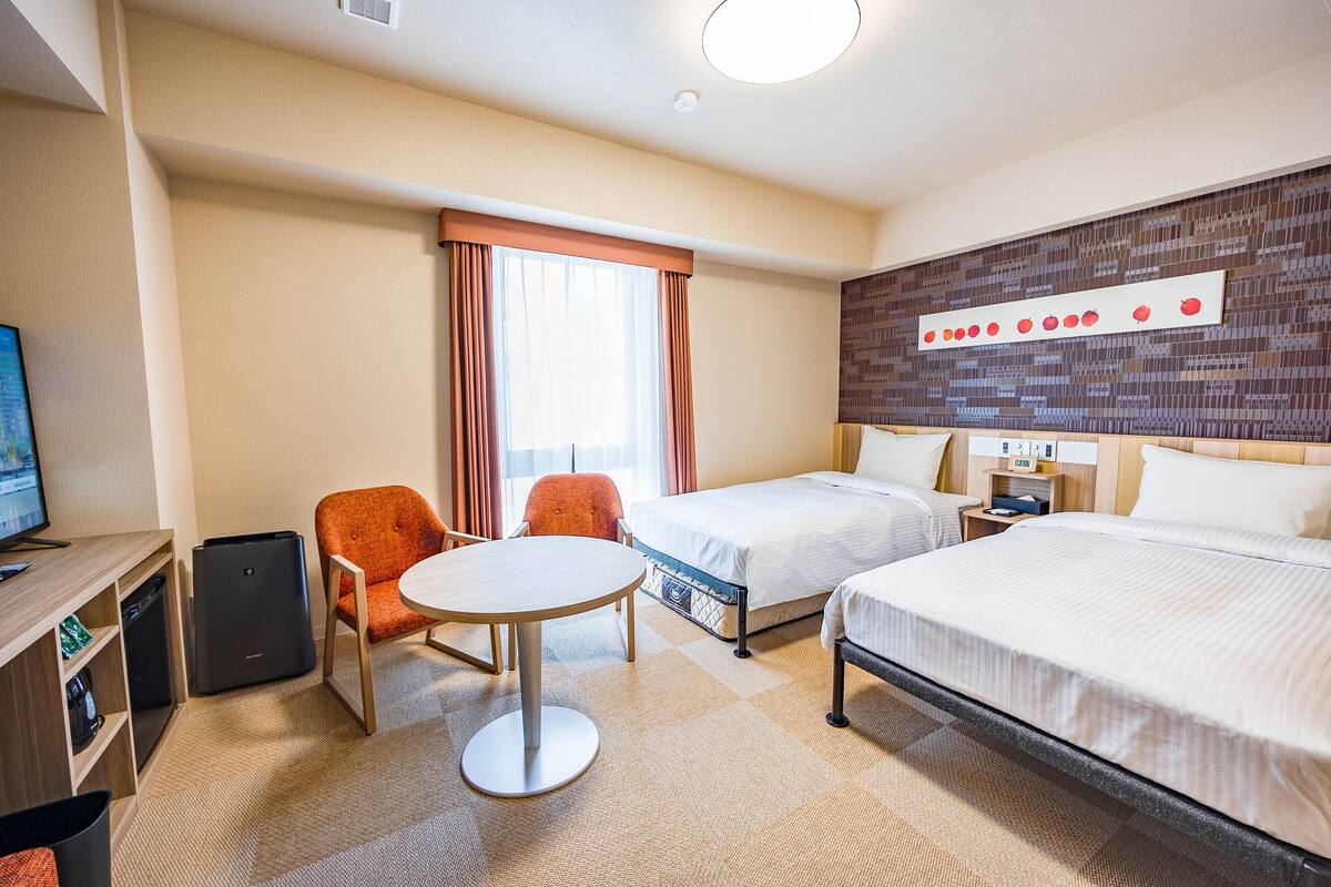 A hotel room with two beds, a desk with a computer, a chair, a table, and a wall with a decorative pattern.