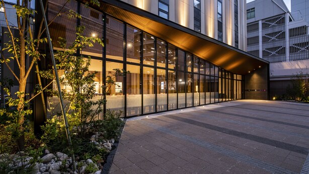 Un edificio moderno con amplios ventanales, techo curvo y un acceso pavimentado con rocas y plantas.