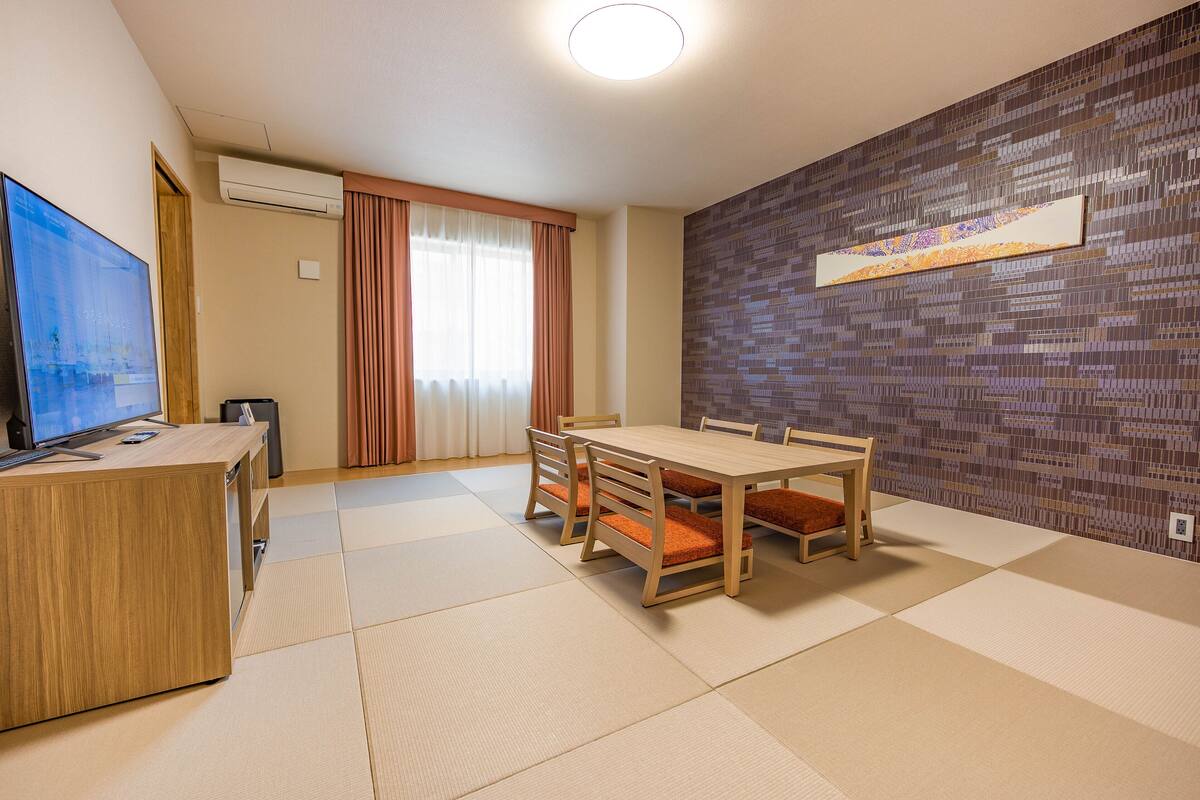 A room with a wooden desk, a television, a dining table with chairs, a patterned wall, and a sliding door with curtains.