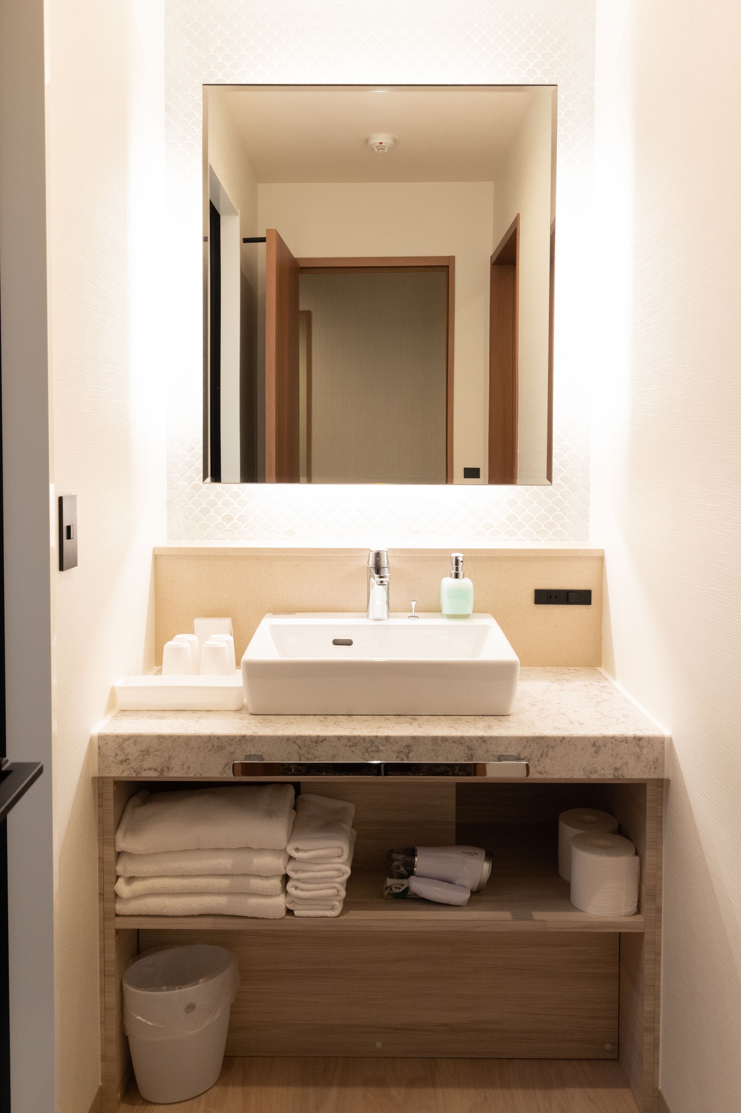 A bathroom with a white sink, a mirror above it, and a wooden shelf below holding towels and toiletries.