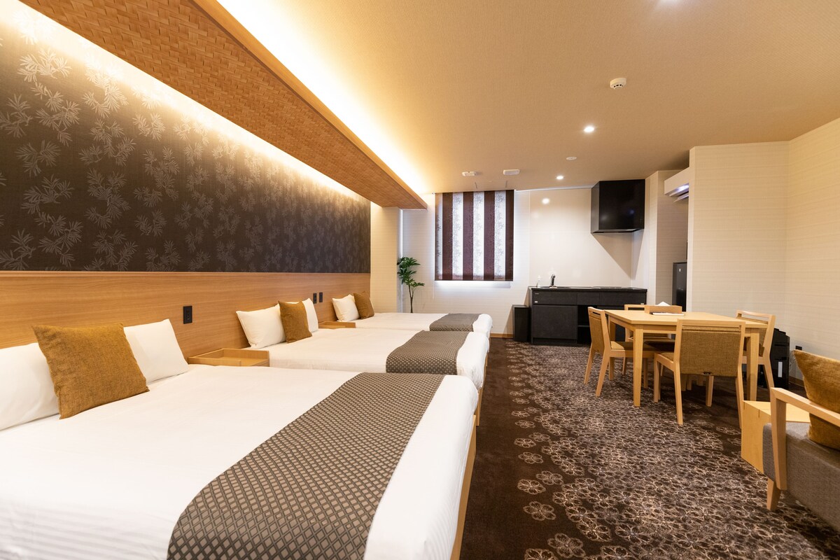 Ein Hotelzimmer mit drei Betten, einem Esstisch, einem Fernseher und einer Pflanze.