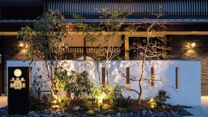 Ein Hotel mit traditioneller japanischer Architektur, mit einem Kiesweg, einem gepflegten Garten mit Bäumen und Sträuchern sowie einem Schild mit dem Namen des Hotels.
