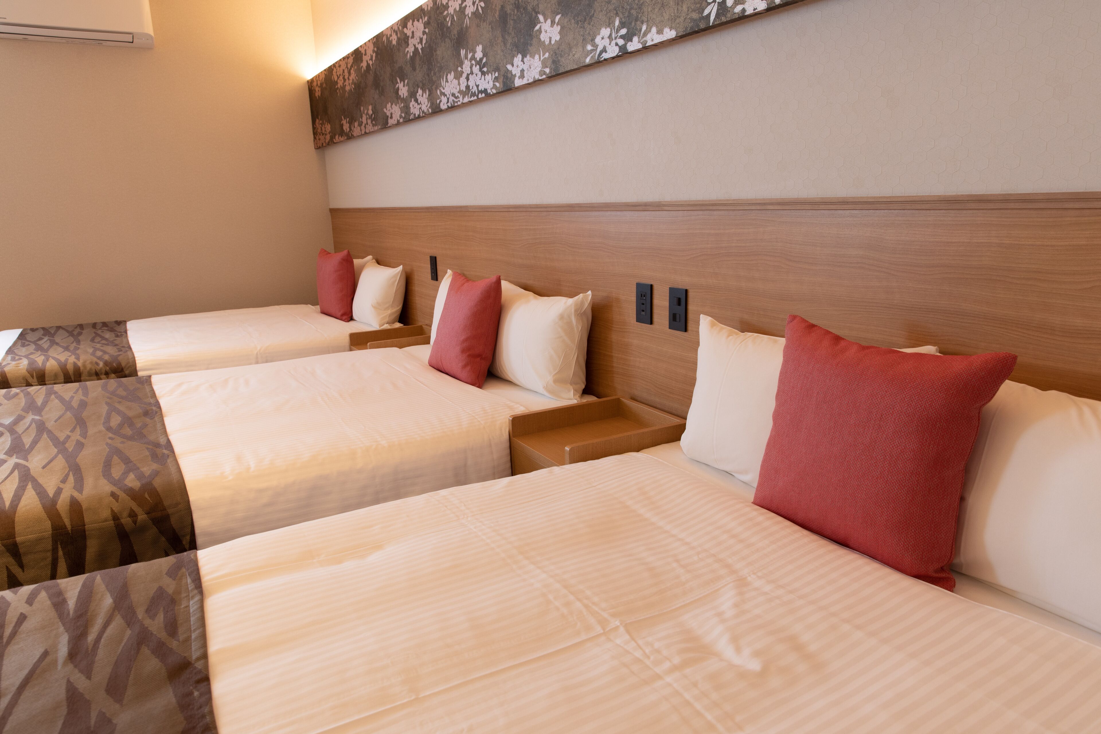 Habitación de hotel con tres camas, cada una con cabecera de madera, mesita de noche y almohada roja.