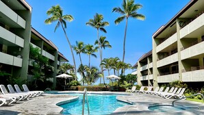 A pool area with lounge chairs and palm trees, surrounded by multi-story buildings. - Peaceful Kona Retreat—Steps to Magic Sands Beach · Ocean View · King Beds · AC! (Kailua-Kona)