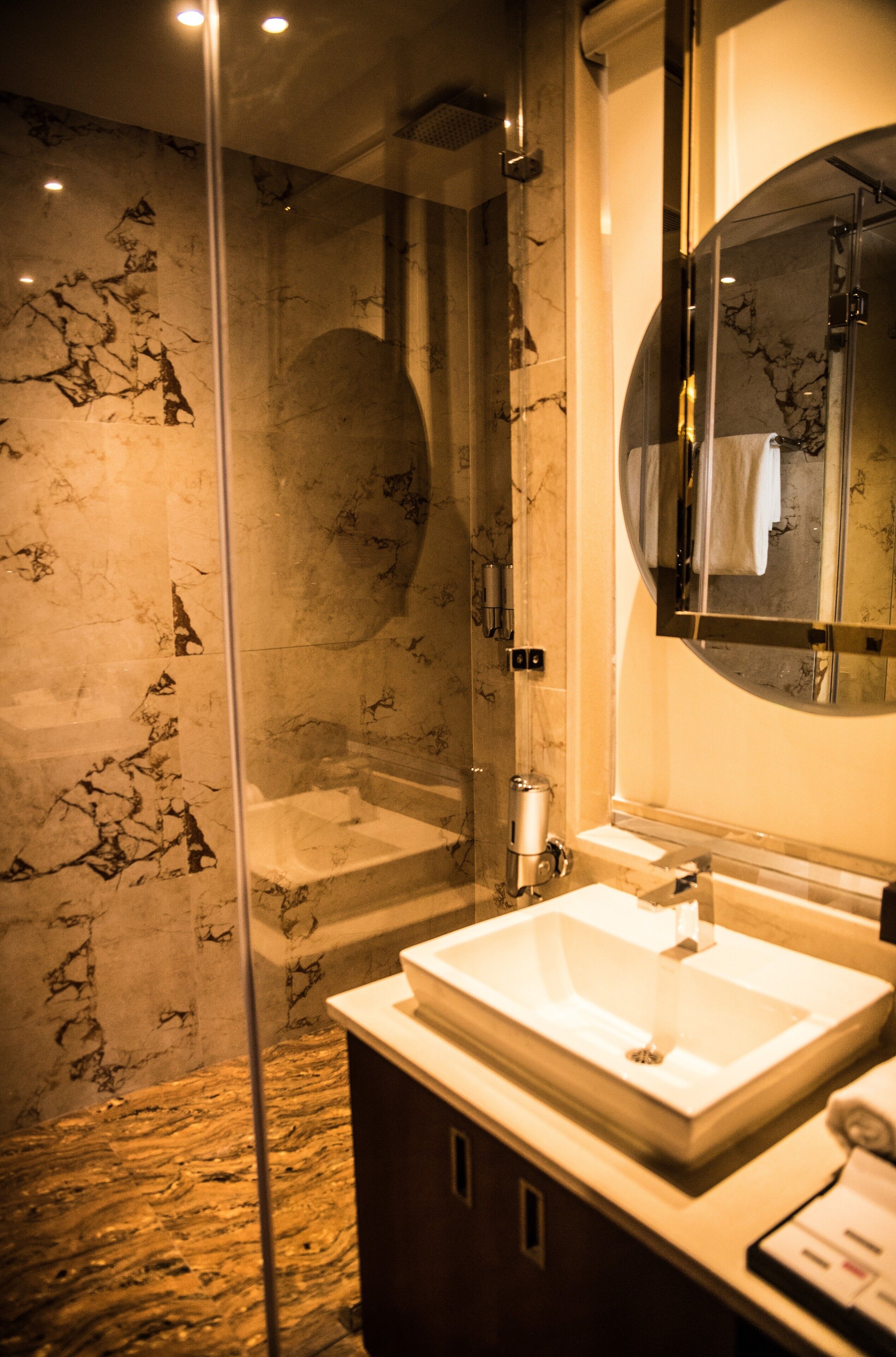 A modern bathroom with a glass-enclosed shower, a marble-tiled wall, and a large mirror above the sink.