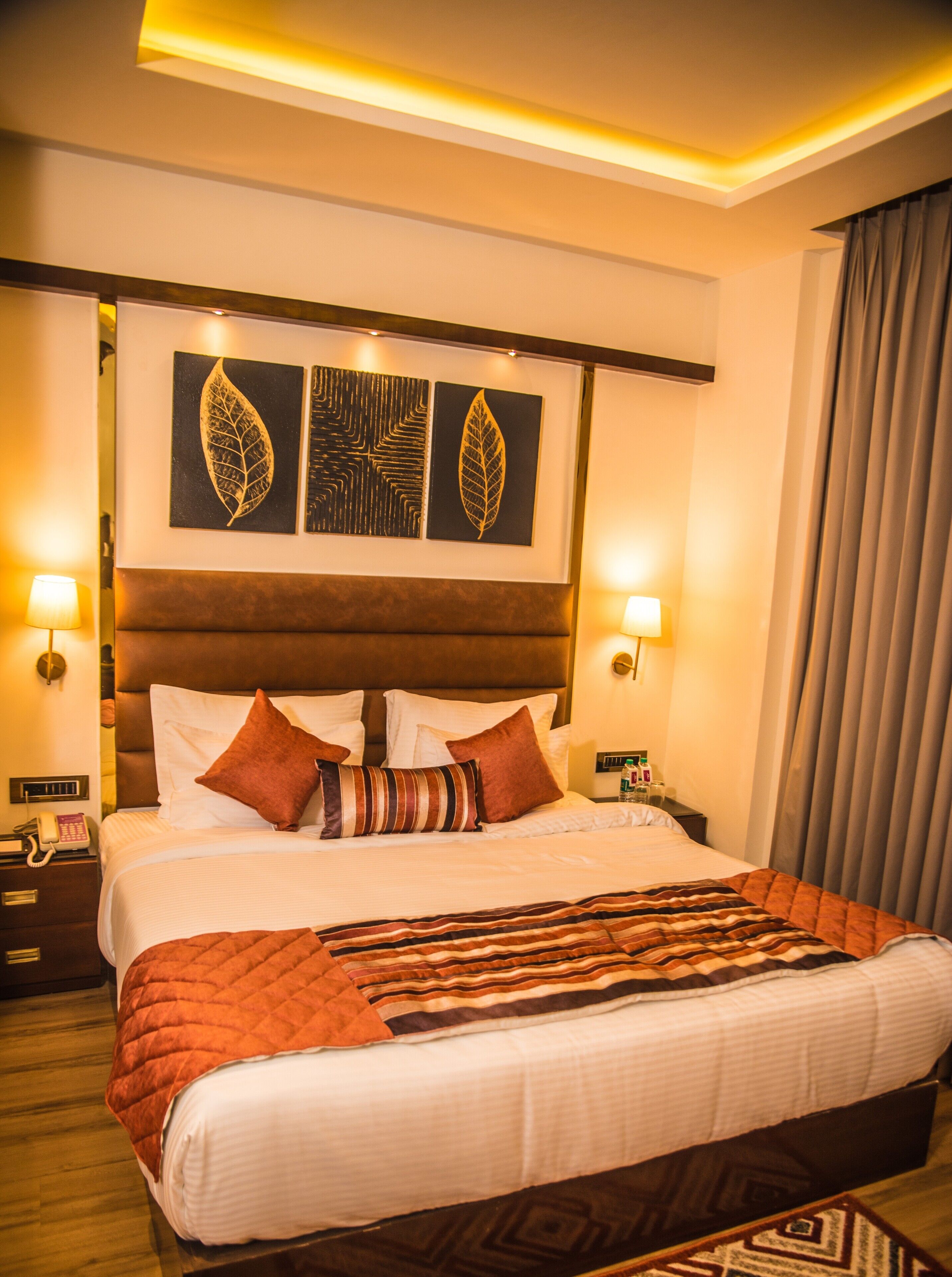 A hotel room with a large bed, two wall-mounted lamps, a headboard with leaf-patterned artwork, and a bedside table with a phone and bottle.