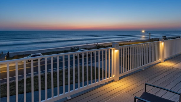 Une terrasse en bois avec une balustrade blanche donnant sur une plage au coucher du soleil.