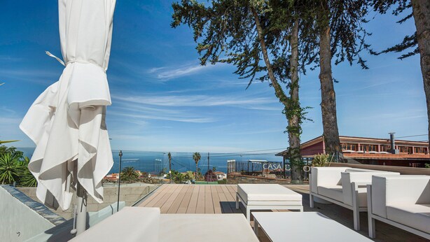 An outdoor seating area with white furniture, a white umbrella, and a view of the sea and buildings in the background. - "Loft la Matanza" with Sea View, Mountain View, Pool, Terrace & Garden (La Matanza)