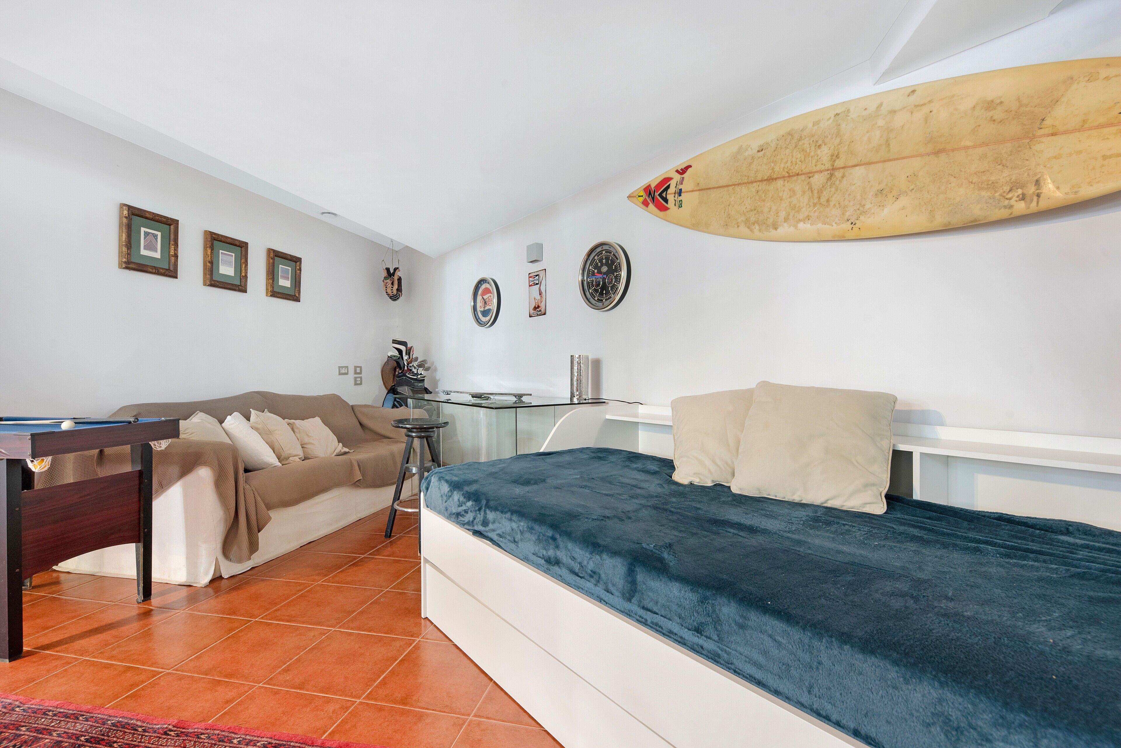 A room with a bed, a pool table, a sofa, and a surfboard on the wall.