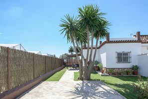 Un patio trasero cercado con un sendero de hormigón, palmeras y un edificio blanco con ventana y pared de piedra.