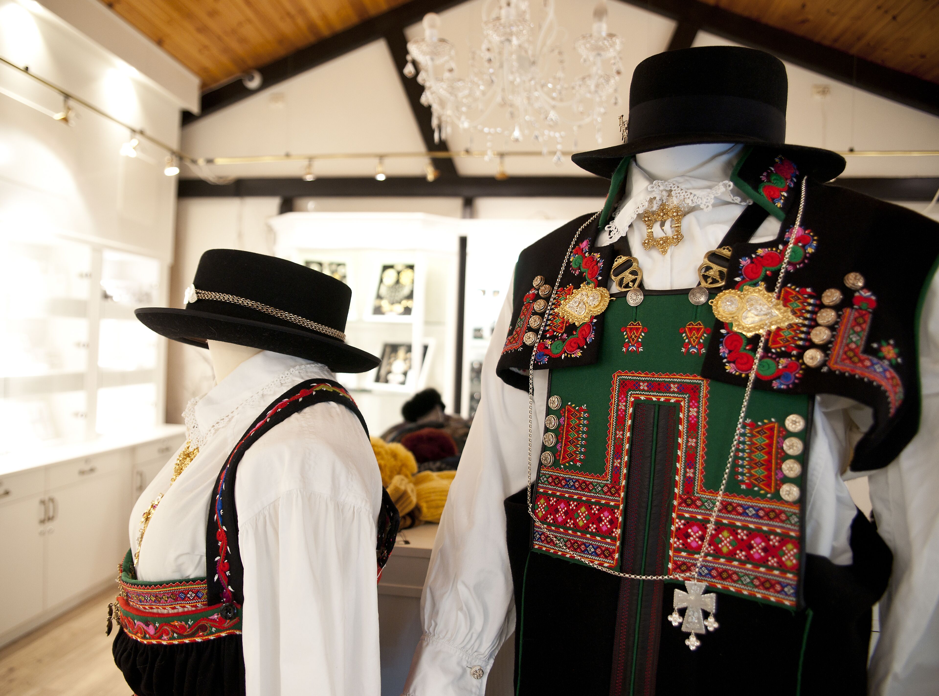 Maniquíes vestidos con atuendo tradicional con bordados intrincados y accesorios, exhibidos en una sala bien iluminada con techo de madera y candelabros.