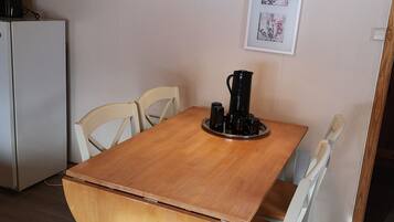 Una mesa de comedor de madera con cuatro sillas, una lámpara colgante roja, una cafetera negra y un refrigerador al fondo.
