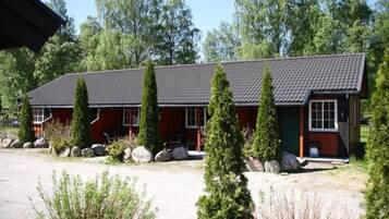 A two-story wooden cabin with a dark roof, surrounded by greenery and rocks.