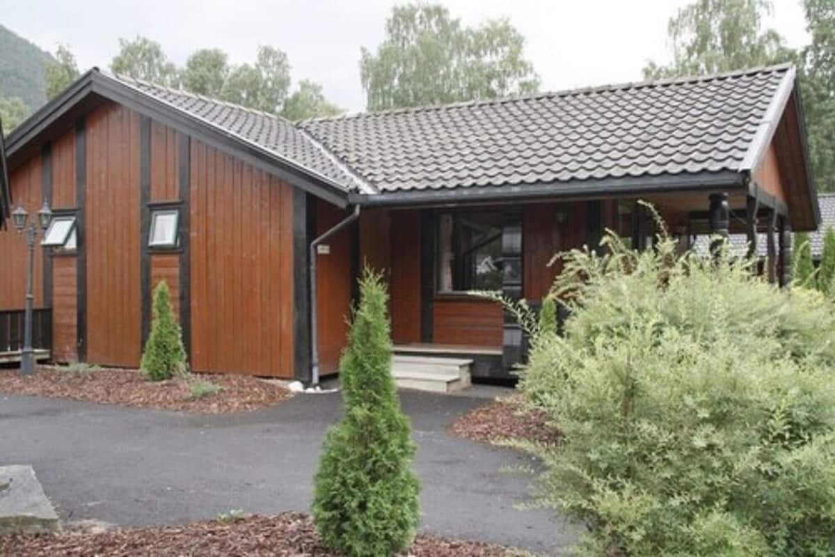 Eine Holzhütte mit einem Ziegeldach, einer Veranda und einem kleinen Vorgarten mit Sträuchern.