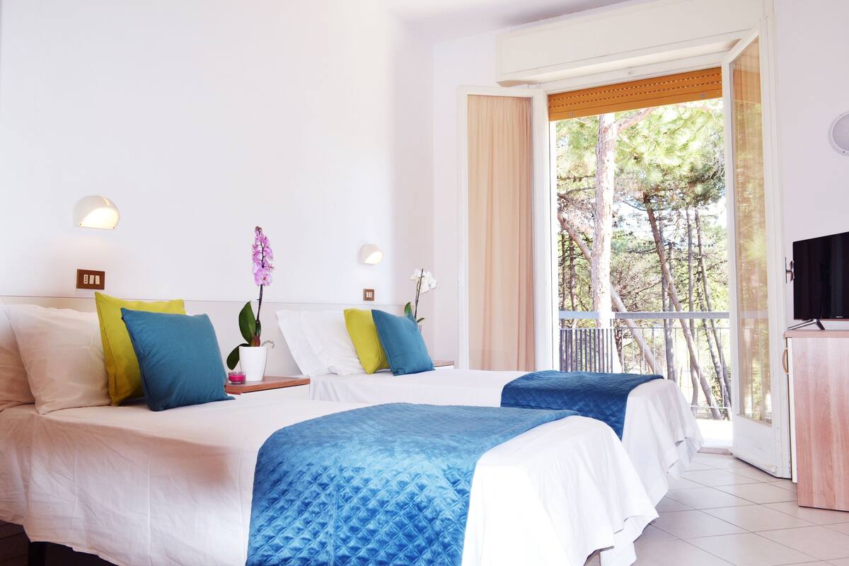 A hotel room with two beds, a balcony with a view of trees, and a TV on a wooden cabinet.