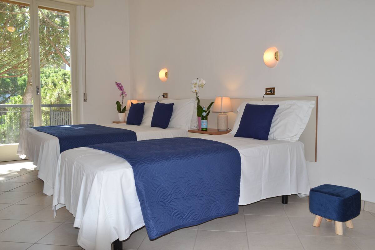 A hotel room with two beds, each with a white and blue bedspread, a small bedside table with a lamp, and a potted orchid.