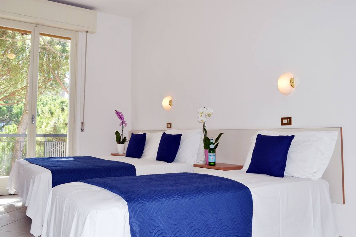 A hotel room with two beds, white bedding, blue accents, and a balcony with a view of greenery.