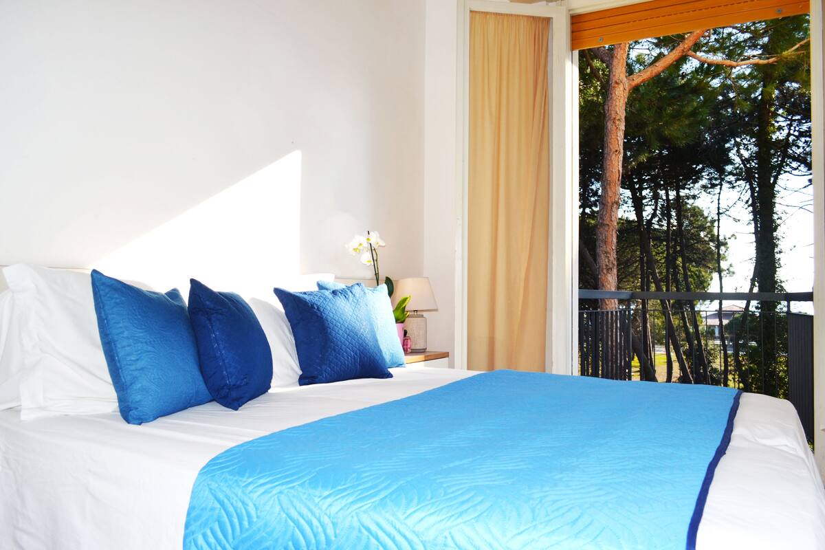 A hotel room with a large bed, blue bedding, a nightstand with a lamp, and a view of trees through a balcony.