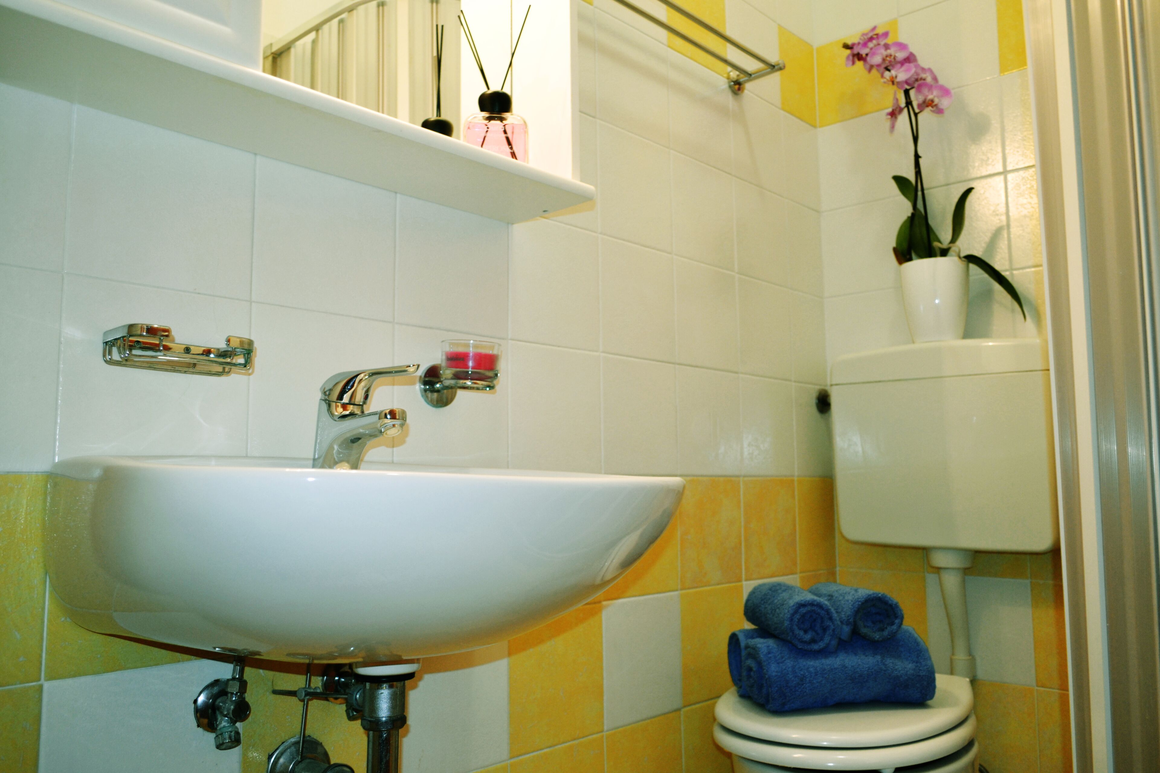 A bathroom with a white sink, yellow and white tiles, a toilet with blue towels, and a potted orchid.