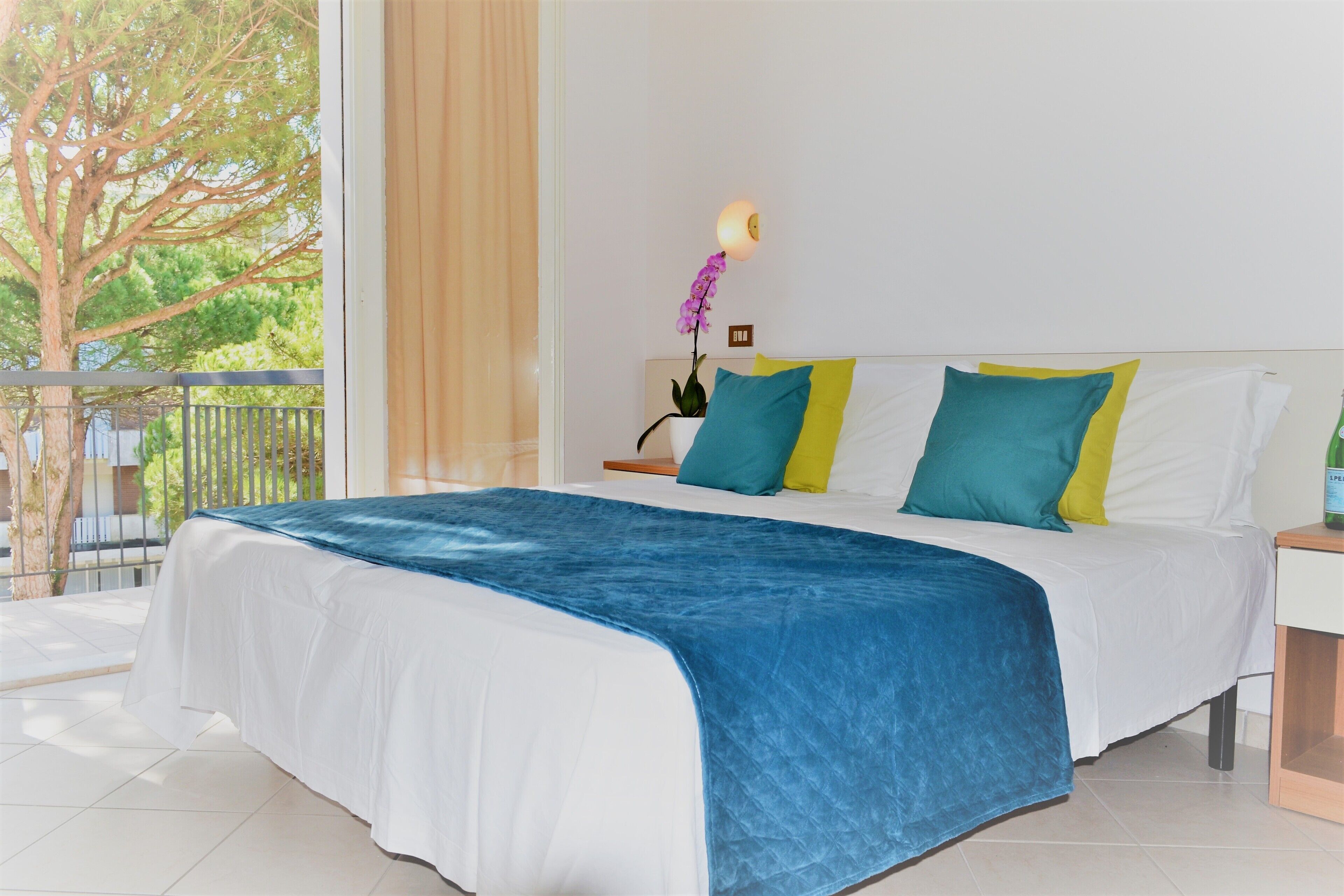 A hotel room with a large bed, a nightstand, a lamp, and a view of a balcony with a tree.