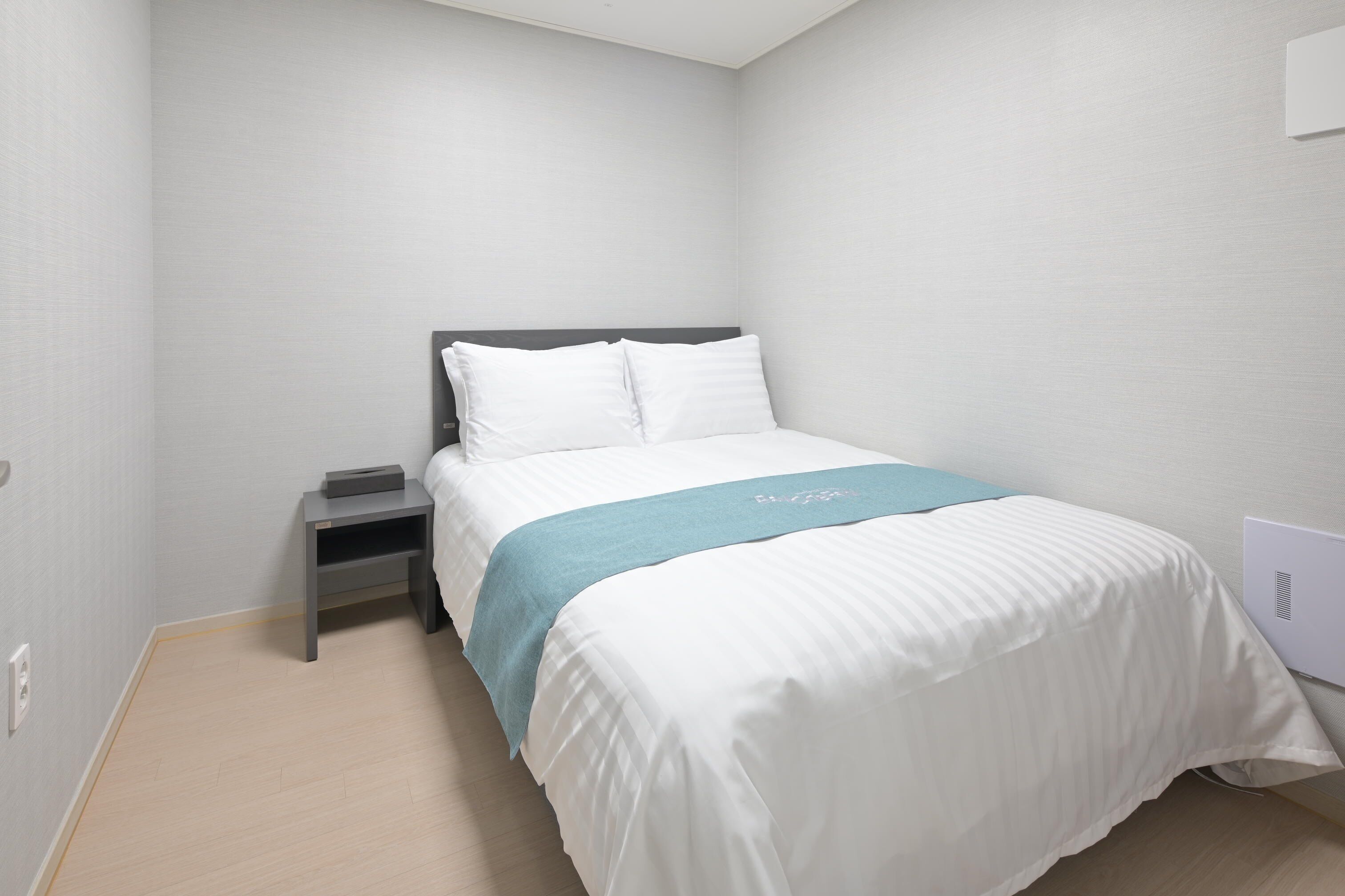 A neatly made bed with white bedding and a blue accent, a bedside table with a lamp, and a wall-mounted power outlet.