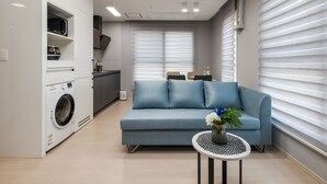Un espacio habitable funcional con lavadora, sofá, una mesita y un florero, además de una ventana con persianas.