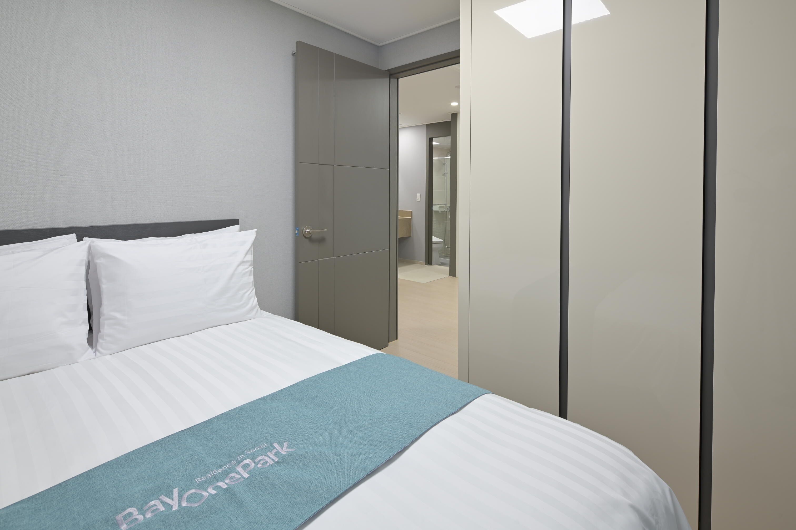 A neatly made bed with white linens and a blue blanket, a grey wardrobe, and a bathroom visible through an open door.