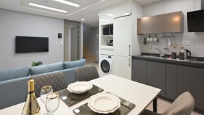 Cocina moderna con horno empotrado, microondas y lavabo. Hay una mesa de comedor con platos y vasos, y un sofá al fondo.