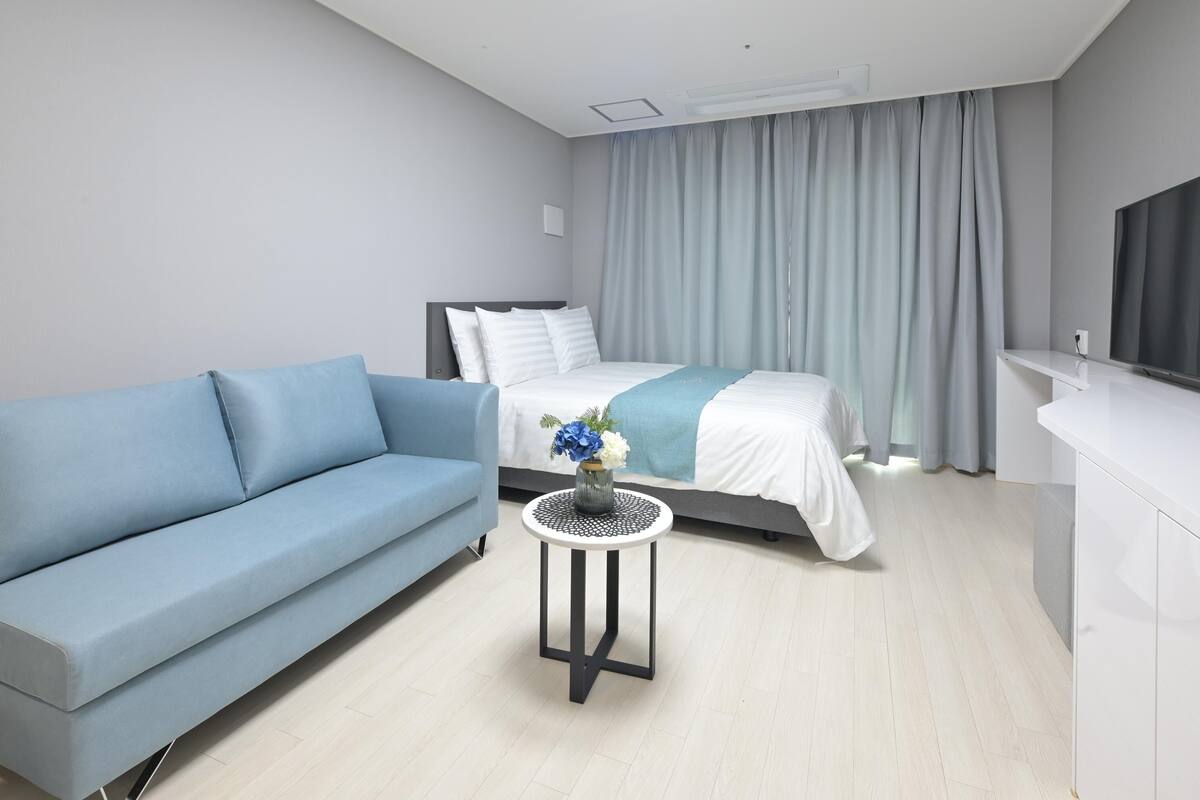 A modern hotel room with a bed, a blue sofa, a small round table with a vase, and a TV on a white cabinet.