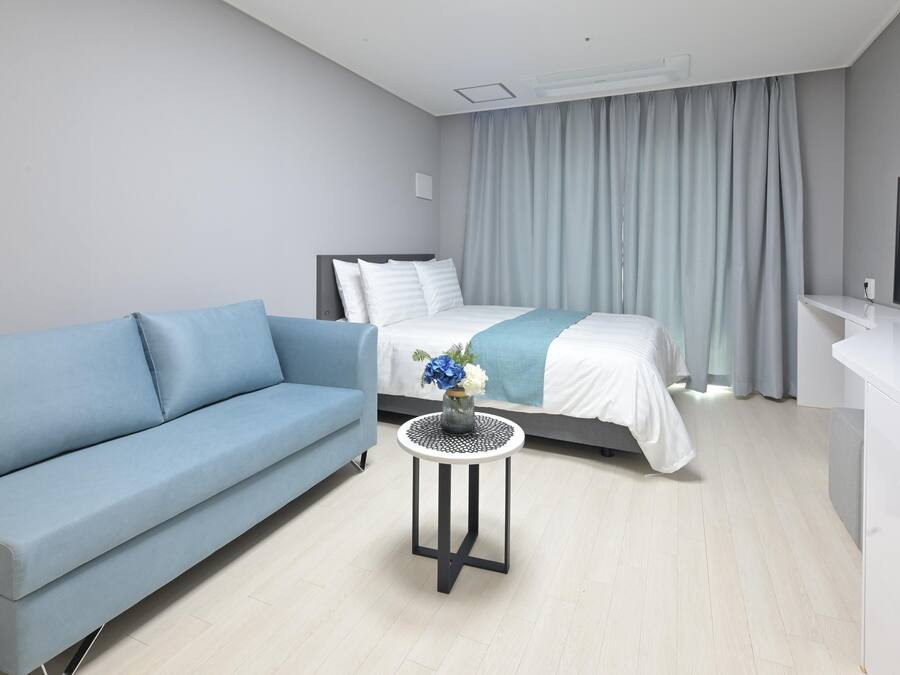 A modern hotel room with a bed, a blue sofa, a small round table with a vase, and a TV on a white cabinet.