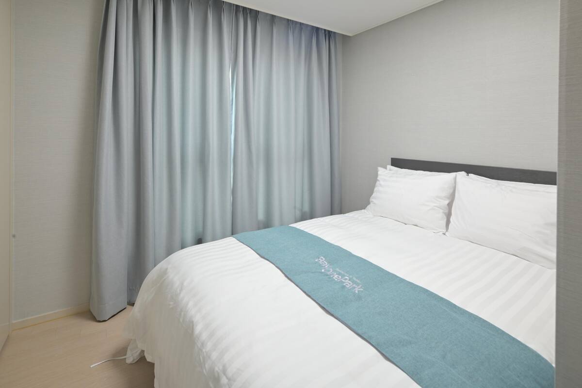 A neatly made bed with white linens and a light blue throw blanket, a grey headboard, and a grey curtain.