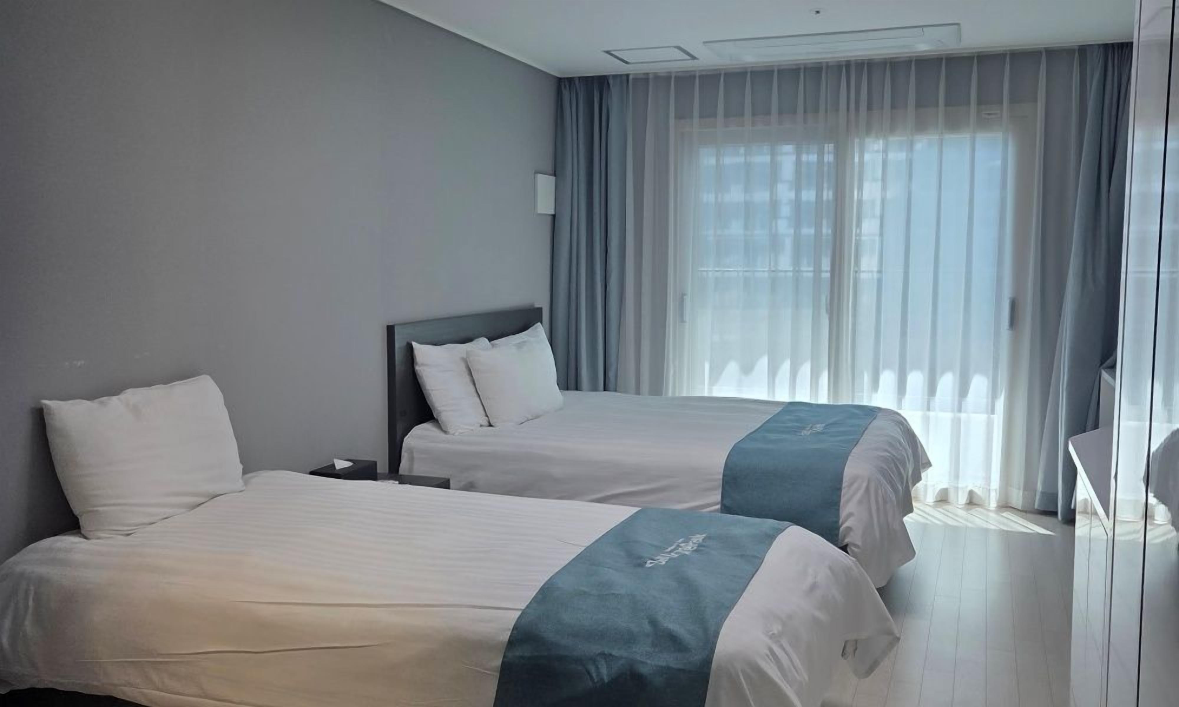 Una habitació d'hotel amb dos llits, una tauleta de nit i una finestra gran amb cortines primes.