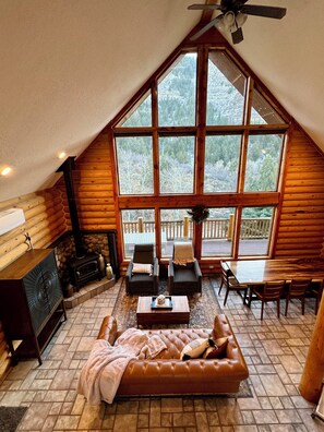 Interior acogedor de una cabaña de madera con un ventanal grande, chimenea, sofá y zona de comedor.