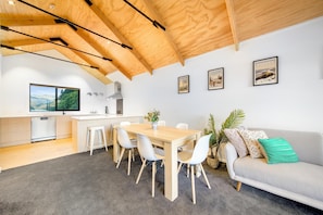 A modern kitchen with a wooden ceiling, a dining area with a wooden table and white chairs, a grey sofa with cushions, and framed pictures on the wall.