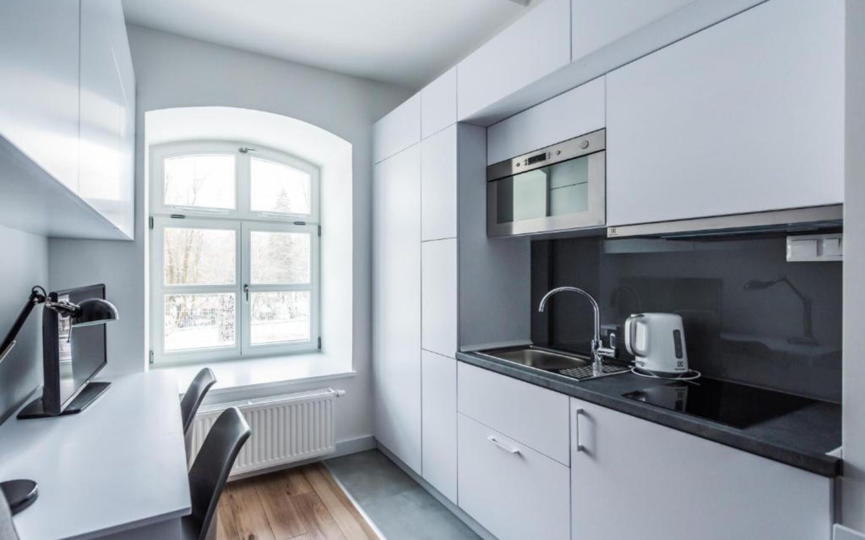 Eine moderne Küche mit weißen Schränken, einer schwarzen Arbeitsplatte, einem eingebauten Backofen und einem Fenster mit Blick auf Bäume.